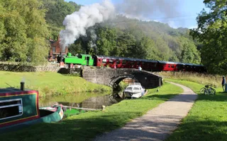 A green and red steam train moves over a bridge above a calm canal with moored boats