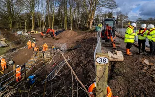Staff in yellow high vis and white hard hats stand gathered round a clipboard, surrounded by work machinery and other staff in orange safety gear as they work.