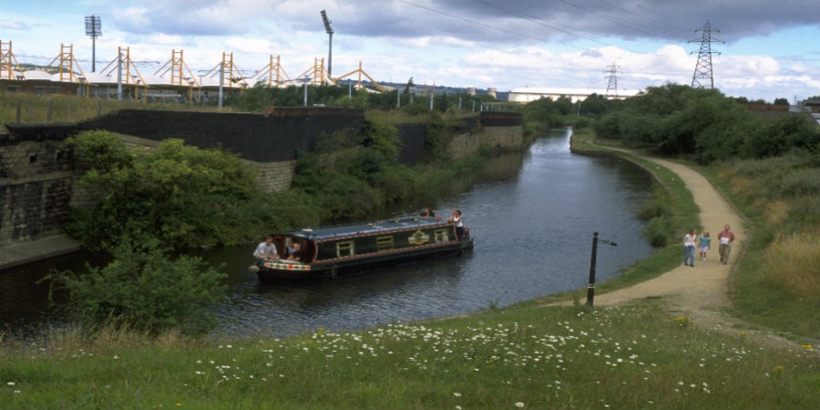 Boat on navigation in front of sports arena, with family walking on towpath