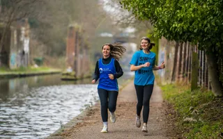 Two women jog along the towpath in an urban area, laughing, wearing 'Canal & River Trust' running vests.
