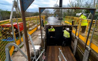 Three men in hi-vis and hard hats work on installing a lock gate along a canal lined with yellow safety barriers.