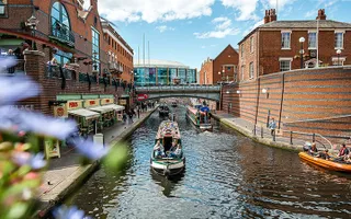 Several narrow boats on the canal, lined by restaurants and onlookers on a sunny day in Birmingham.