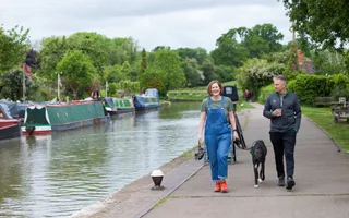 A woman in a blue dungarees holds the lead of a black dog, whilst a man holding a coffee cup walks next to her along the towpath, which is parallel to the canal with moored boats along its edge.