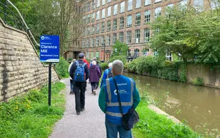 A group of people in coats and jackets walk along a grass-lined towpath away from the camera, with the canal running along their right