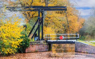 A black, metal lift bridge over the canal on an autumn day, surrounded by golden, yellow and orange leaves.