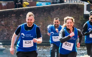 Four adults run along the canal towpath in Birmingham, wearing 'Canal & River Trust' vests and race numbers