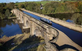 A birds-eye view of a boat travelling over a large beige-stone aqueduct, with a wide river crossing below it
