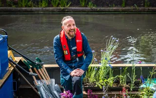 Man in lifejacket sitting in boat filled with flowering plants and spades
