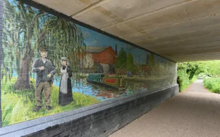 A mural on a bridge wall of a traditional working boat surrounded by an industrial building and two people in period clothing.
