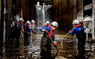 Several colleagues in waterproof protective gear and hard hats stand in the bottom of a lock, sifting through the water.