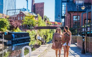 Two women in summer clothing and sunglasses walk along the towpath towards the camera smiling, with the canal to the right