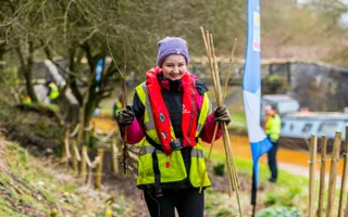 Young volunteer in high vis and life jacket carrying bamboo sticks along the canal towpath