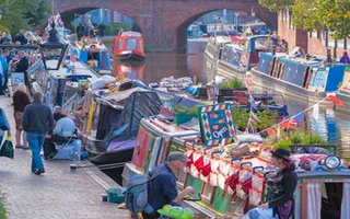 Crowds walk along the towpath, passing many boats covered in bunting selling various items.