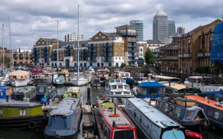 Lots of narrowboats, widebeams, and cruisers moored in a busy basin, surrounded by blocks of flats and Canary Wharf visible in the distance.