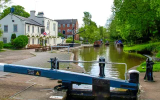 A single lock gate in front of a pound filled with moored boats, a canalside pub with outdoor seating and an old winch.