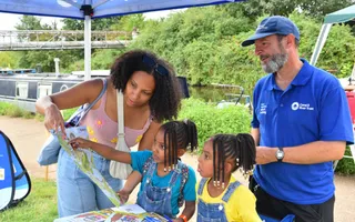 A mother and two young children look at a booklet alongside a Canal & River Trust volunteer, under a gazebo next to the canal