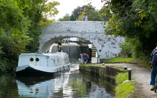 Widebeam boat leaves a lock while a cyclist passes on the towpath and onlookers watch from a bridge