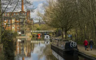 Walkers and moored boats on an autumnal canal, with industrial buildings in the background