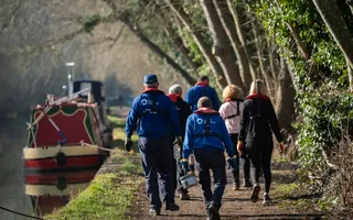 Group of volunteers walk passed a boat along the towpath in life jackets carrying painting equipment