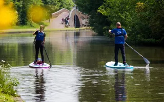 Two people wearing Canal & River Trust branded clothing stand up paddle board along the canal