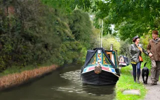 Two adults walk hand in hand with their dog along the canal towpath, passing a moored boat