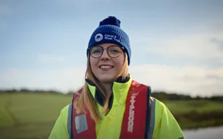 Head and shoulders image of a smiling canal engineer wearing a branded beanie hat, high vis jacket and life jacket in front of wintery canal scene