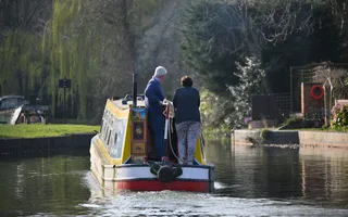 Couple driving a narrowboat along the canal on a sunny day, surrounded by trees