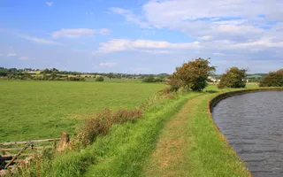 Grassy towpath next to an empty field and canal with blue skies