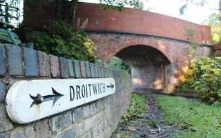 A sign on a brick wall reads 'Droitwich' and has black arrows pointing towards a red bricked bridge.