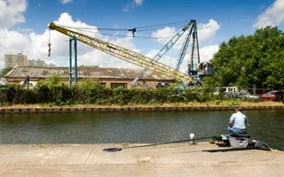 A man sits with his back to camera on a concrete towpath, holding a fishing rod and overlooking the River Lea with tall cranes in the background.