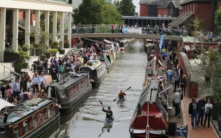 Two people kayak passed moored narrowboats with crowds of people on the towpath and a pedestrian bridge over the canal in a town centre.