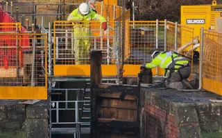 Two workmen in hard hats and hi-vis clothing tend to repairing a lock gate surrounded by yellow safety barriers.