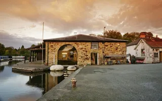 A stone building with a large doorway and a dry dock inside sits by the canal at sunset.