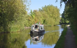 A boat cruises towards the camera in the middle of the canal, which is lined with lush green trees.