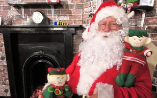 Santa sitting on a chair with toy elves on his lap and shoulder