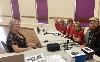 4 young people with 2 adults sitting at a desk with recording equipment inront of them.