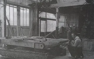 A black and white photo shows a man in period garb drilling into a new lock gate in a large workshop.