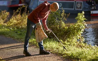 Man litter picking on sunny day next to canal. Carrying bag for rubbish, wearing gloves and carrying litter picker