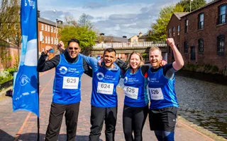 Four charity fun runners celebrating by the canal next to a blue branded flag