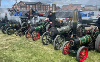 Miniature steam vehicals in a line with people standing behind them.