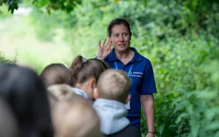 A woman in a blue Canal & River Trust t-shirt stands amongst foliage, holding her hand to her ear to listen for wildlife, whilst children line up in front of her.