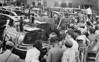 Black and white photo of a festival with people in the foreground standing infront of a line of boats