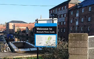 A Canal & River Trust sign saying 'Welcome to Newark Town Lock' in front of a canal lock and old warehouses