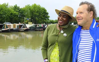 Two people pose in front of moored narrowboats in a marina