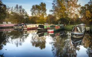 Six boats moored side by side close to a grassy bank, with trees and sky reflected in the water in front