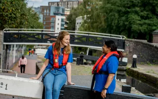 Two female colleagues in life jackets laugh as they sit on the lock beam.