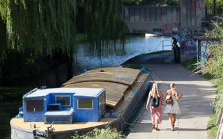 Two women in summery clothing walk along the towpath past a widebeam work boat.