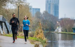Two runners smile as they job along the towpath, with the Birmingham cityscape behind them.