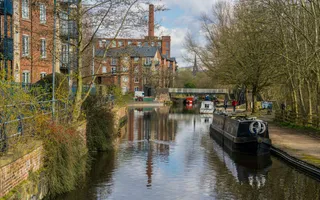 Boats moored along a tree-lined canal with residential buildings on one side and a large brick chimney in the background.