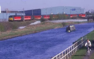 A boat moves along the canal in an industrial setting with a cyclist on the towpath and a train on the opposite side.